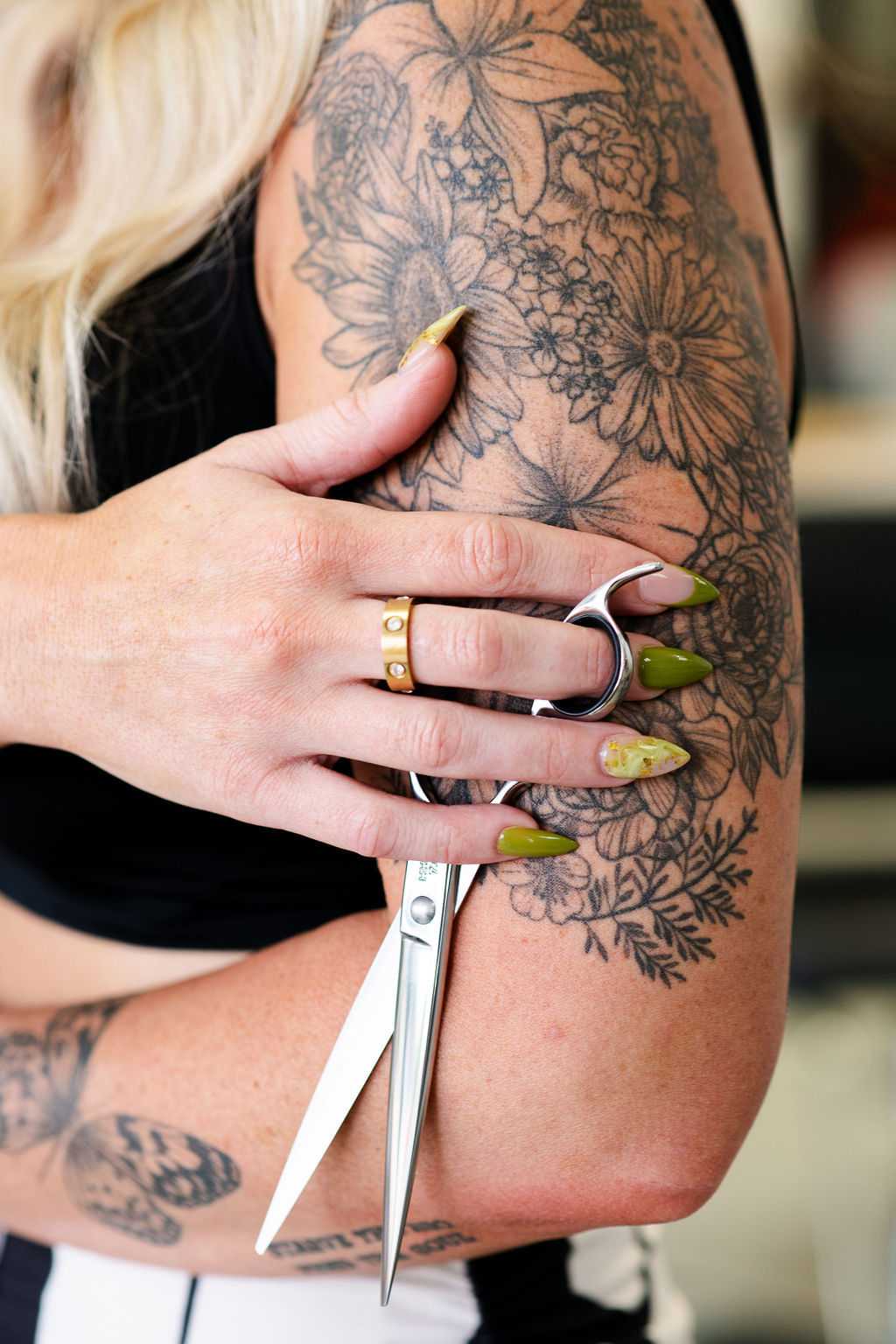Person with floral tattoos holds hairstyle scissors, showcasing intricate nail designs.